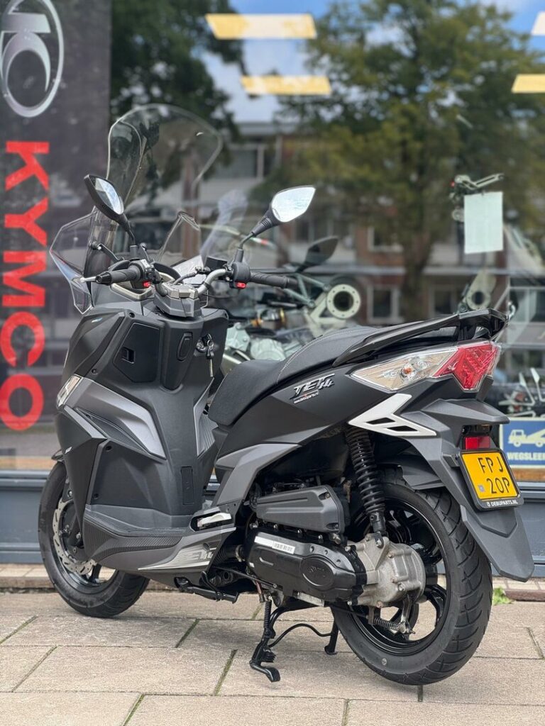 Grijze Kymco-scooter met hoog windscherm, op standaard geparkeerd voor een showroom.