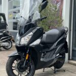 Zwarte maxi-scooter met hoog windscherm, geparkeerd voor een werkplaats