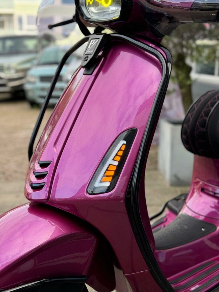 Close-up van de voorkant van een metallic roze scooter met oranje knipperlicht.