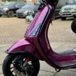 Close-up van een magenta scooter met zwarte details, voorwiel en koplamp, geparkeerd op straat.