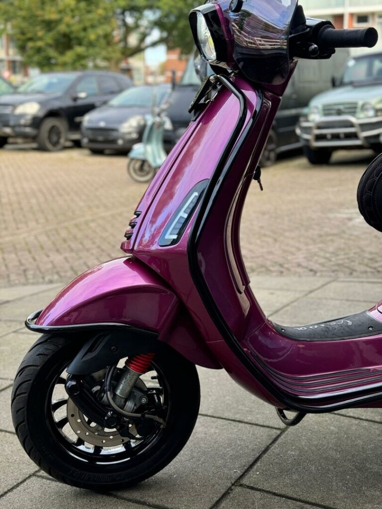 Close-up van een magenta scooter met zwarte details, voorwiel en koplamp, geparkeerd op straat.