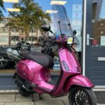 Fuchsia Vespa-scooter met hoog windscherm, geparkeerd voor een winkelpui.