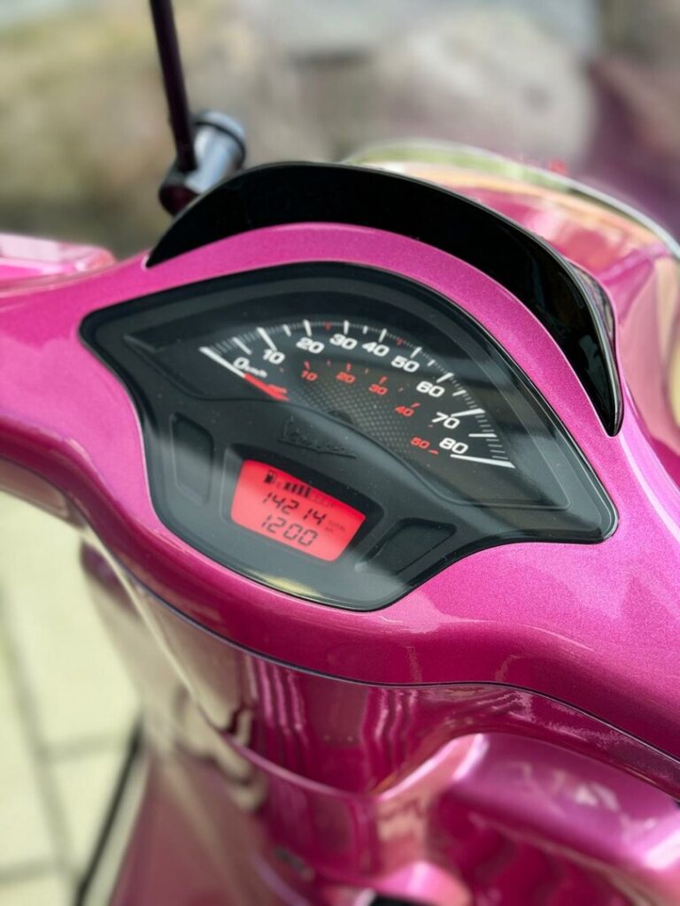 Close-up van de snelheidsmeter en het digitale display op een metallic roze scooter.