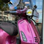 Close-up van een metallic roze Vespa scooter met windscherm en koplamp voor een winkelraam.