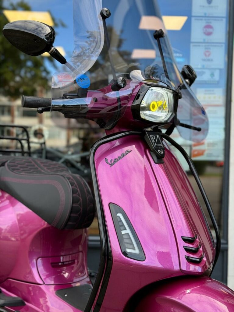 Close-up van een metallic roze Vespa scooter met windscherm en koplamp voor een winkelraam.