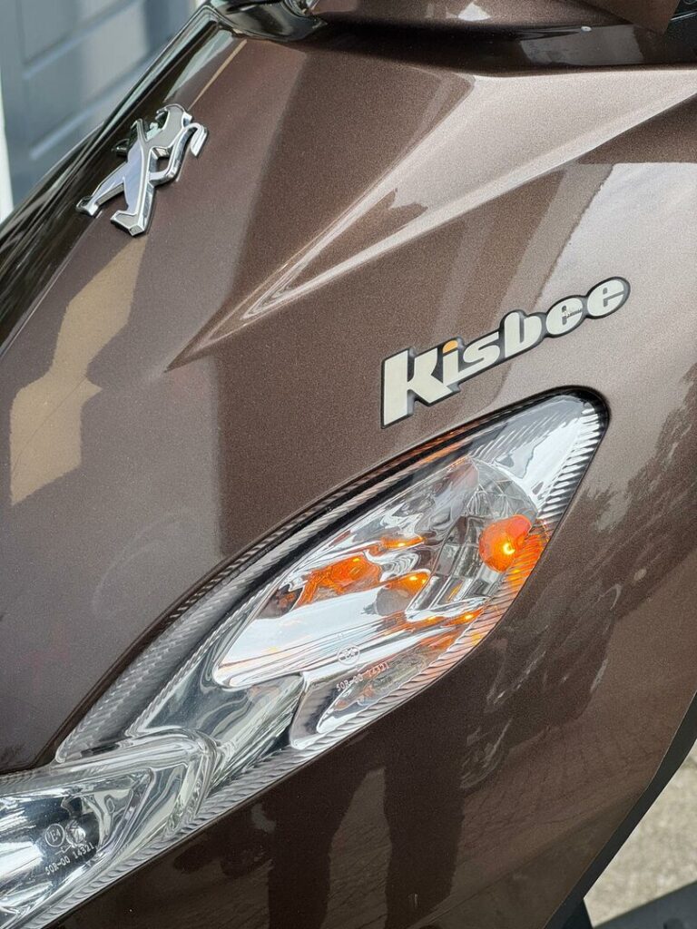 Close-up van de voorkant van een bruine Peugeot Kisbee-scooter met brandend knipperlicht.