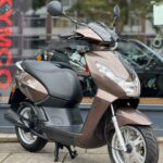 Bruine Peugeot Kisbee scooter met spiegels, op standaard geparkeerd voor een showroom.