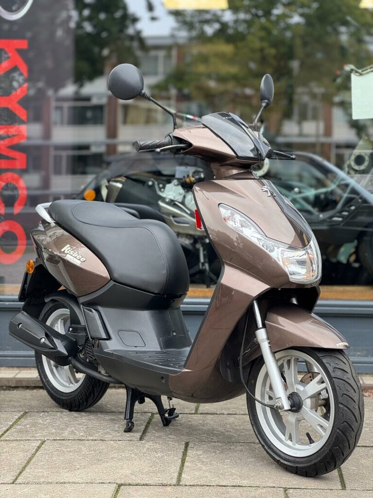 Bruine Peugeot Kisbee scooter met spiegels, op standaard geparkeerd voor een showroom.