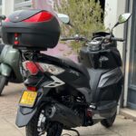 Zwarte scooter met topkoffer en gele kentekenplaat, achteraanzicht, geparkeerd voor een winkel.