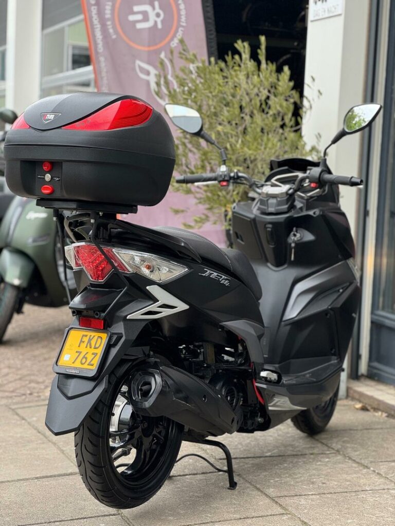 Zwarte scooter met topkoffer en gele kentekenplaat, achteraanzicht, geparkeerd voor een winkel.