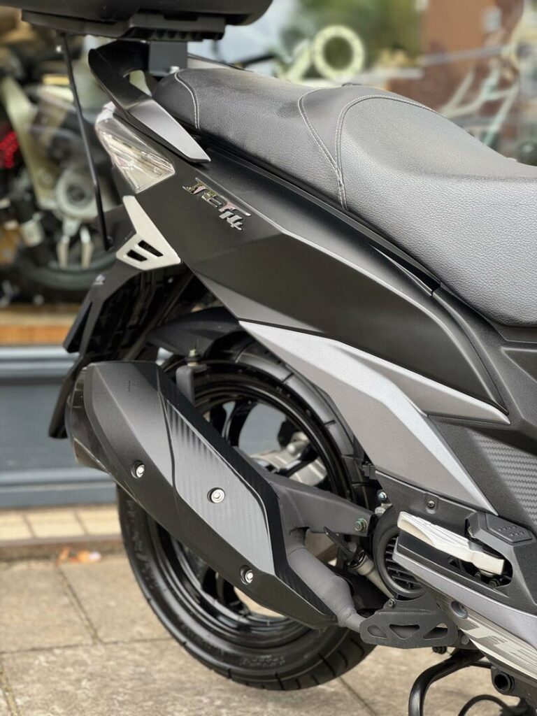 Close-up van de achterzijde van een zwarte scooter met zadel, uitlaat en achterwiel.