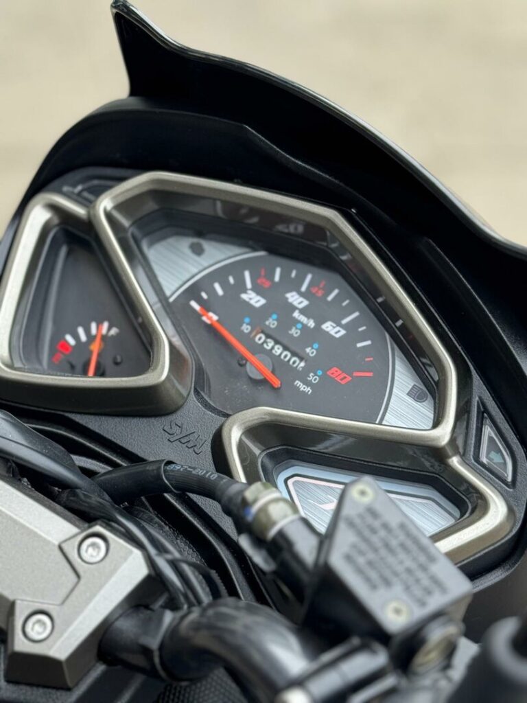 Close-up van het dashboard van een SYM-motor, met analoge snelheidsmeter en brandstofmeter.