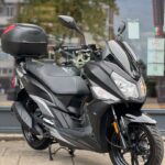 Zwarte motorscooter met topkoffer geparkeerd voor een winkelraam op de stoep.