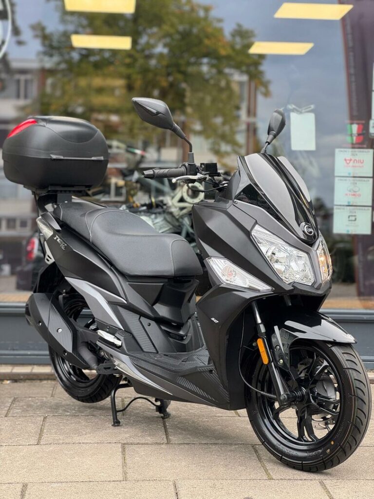 Zwarte motorscooter met topkoffer geparkeerd voor een winkelraam op de stoep.