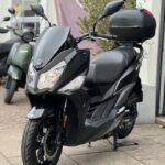 Zwarte SYM-scooter met topkoffer geparkeerd voor een scooterwinkel, schuin van voren gezien.