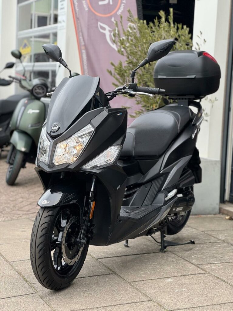 Zwarte SYM-scooter met topkoffer geparkeerd voor een scooterwinkel, schuin van voren gezien.