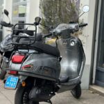 Grijze Riva-scooter met blauw Nederlands kenteken, geparkeerd voor een winkelpui.