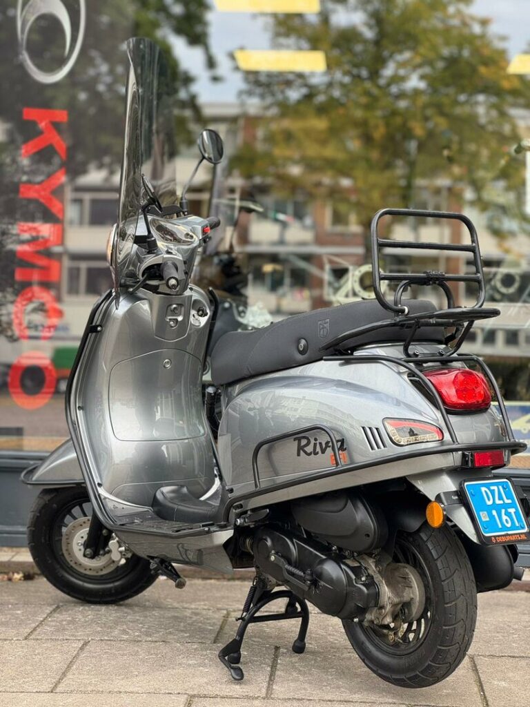 Zilveren retro-scooter met hoog windscherm en bagagedrager, geparkeerd voor een etalage.