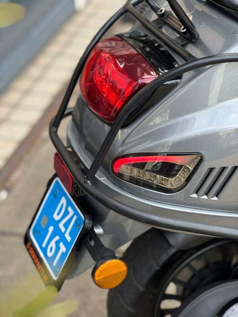 Achterkant van een grijze scooter met bagagerek, achterlicht en blauwe kentekenplaat.