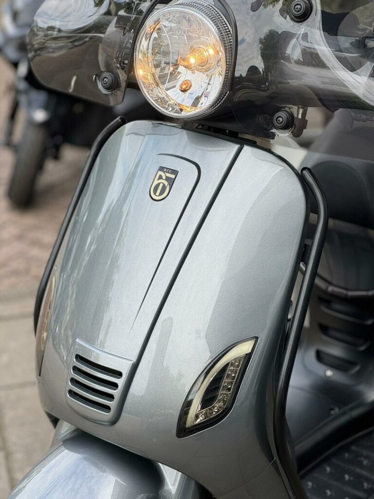 Close-up van de voorkant van een grijze scooter met brandende koplamp en windscherm.