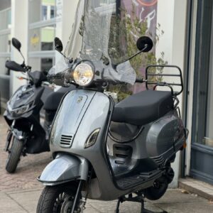 Grijze scooter met hoog windscherm en bagagerek, geparkeerd voor een werkplaats.