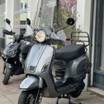 Grijze retro-scooter met hoog windscherm, geparkeerd voor een winkel.