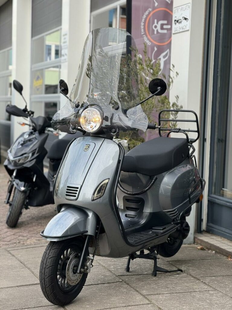 Grijze retro-scooter met hoog windscherm, geparkeerd voor een winkel.