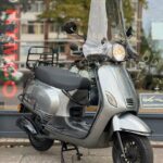 Grijze scooter met hoog windscherm en bagagedrager geparkeerd voor een winkelraam.