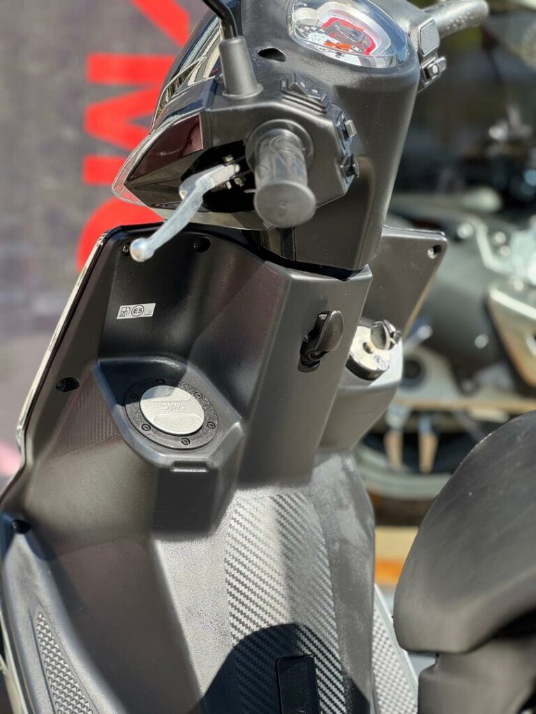 Close-up van het stuur, dashboard, remhendel en voetplaat van een zwarte scooter in zonlicht.