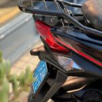 Close-up van de achterkant van een zwarte scooter met bagagerek, rode achterlamp en blauwe kentekenplaat.