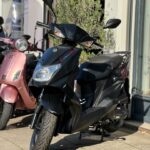 Zwarte SYM Orbit III scooter geparkeerd op trottoir voor winkel, zonlicht, vooraanzicht.