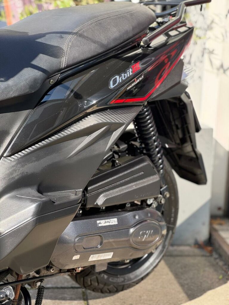 Close-up van de achterzijde van een zwarte scooter met zadel, vering en motorbehuizing.