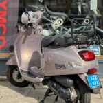 Zandkleurige AGM VX50s scooter op standaard, buiten voor een showroom.