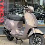 Beige glanzende retro scooter met bagagedrager, geparkeerd voor een motorwinkel.