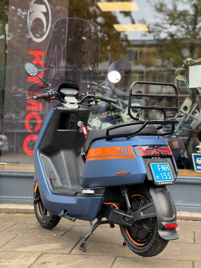 Blauw-oranje elektrische scooter met windscherm en bagagerek, geparkeerd voor een winkel.