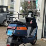 Achteraanzicht van een blauw-oranje scooter met bagagedrager, geparkeerd bij een garage