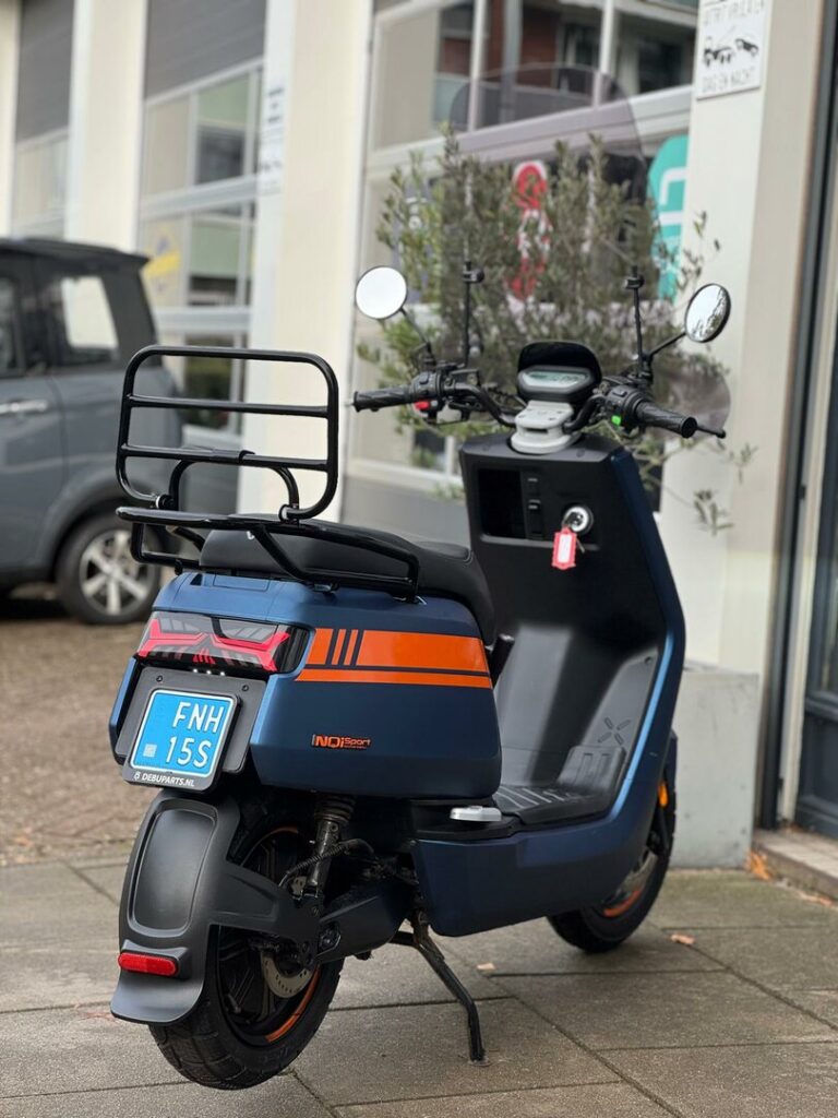 Achteraanzicht van een blauw-oranje scooter met bagagedrager, geparkeerd bij een garage