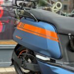 Close-up van een matblauwe scooter met oranje striping, achterrek en achterwiel met schijfrem