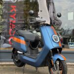 Blauw-oranje elektrische scooter met hoog windscherm, geparkeerd voor een winkel