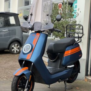 Blauw-oranje NIU elektrische scooter met hoog windscherm, geparkeerd op de stoep voor een werkplaats.