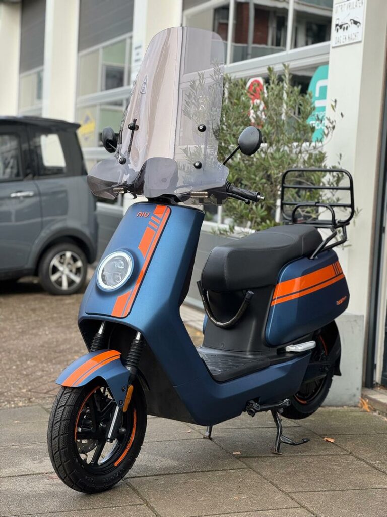 Blauw-oranje elektrische scooter met hoog windscherm, geparkeerd op het trottoir voor een garage.