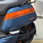 Close-up van de zijkant van een matte blauwe scooter met oranje striping, zadel en bagagedrager.