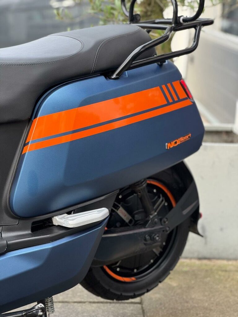 Close-up van de zijkant van een matte blauwe scooter met oranje striping, zadel en bagagedrager.
