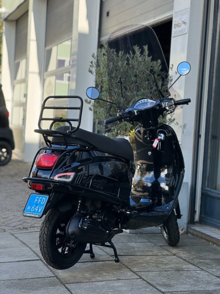 Zwarte retro-scooter met bagagerek en blauwe kentekenplaat, geparkeerd voor een werkplaats