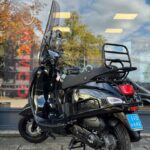 Zwarte scooter met windscherm en bagagedrager, geparkeerd voor een winkelpui.