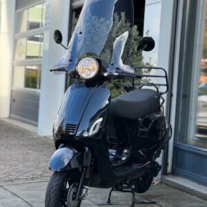 Zwarte scooter met hoog windscherm geparkeerd voor een garage of werkplaats.