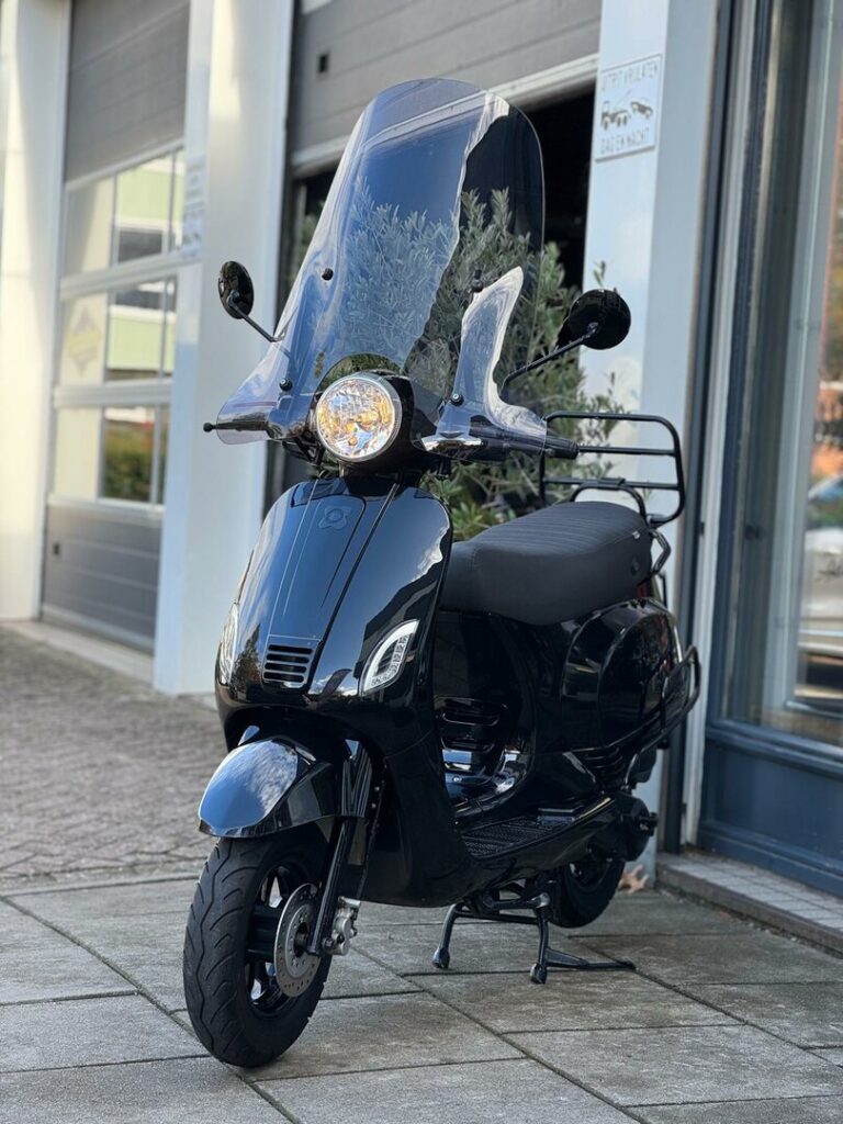 Zwarte scooter met hoog windscherm geparkeerd voor een garage of werkplaats.