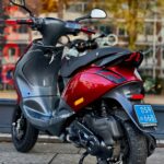 Rode scooter met zwarte accenten en blauwe kentekenplaat, geparkeerd op straat bij gebouwen.