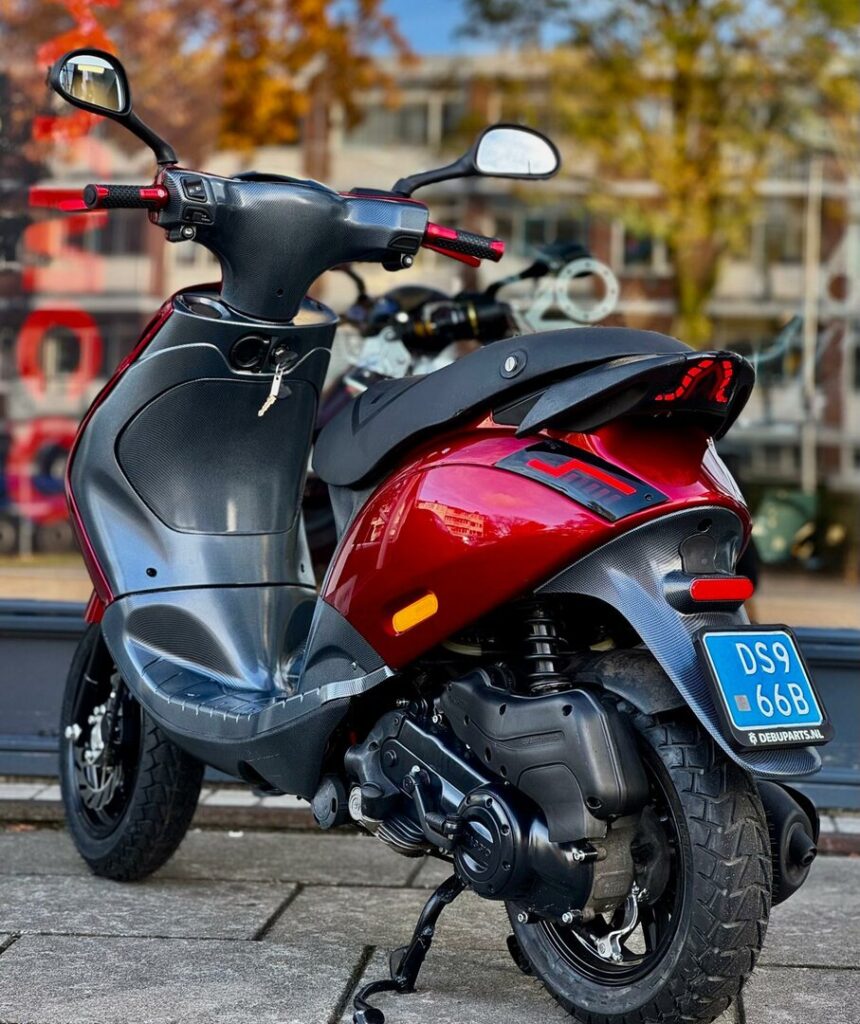 Rode scooter met zwarte accenten en blauwe kentekenplaat, geparkeerd op straat bij gebouwen.