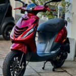 Glanzend rode scooter met zwart zadel geparkeerd op stoep bij gebouw en plantenbak.